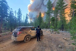 An officer with the Paradise Police Department stands beside a parked patrol vehicle and looks upon towering flames of wildfire.